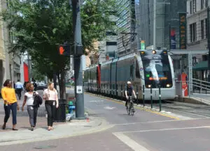 People walking in downtown Houston, while a man passes on his bicycle and the METRO Red line picks up passengers.