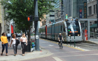 People walking in downtown Houston, while a man passes on his bicycle and the METRO Red line picks up passengers.